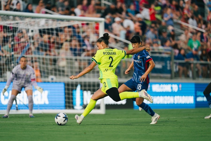Photo of soccer player in yellow kicking toward goal, defended by blue-uniform player, packed crowd