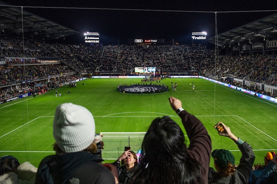 Fans raise arms in packed stadium as teams line up on lit soccer field at night