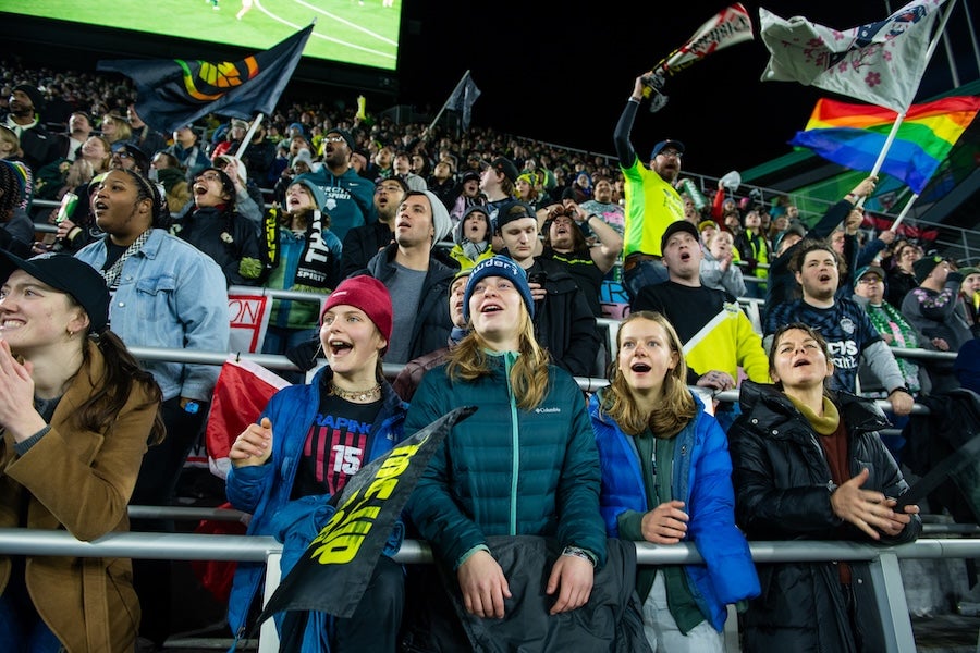 Fans cheer in stadium stands at night, waving colorful flags and scarves