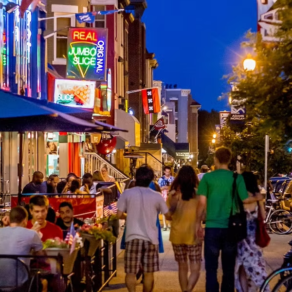 People dining at outdoor tables on a lively city street at night, neon signs glowing