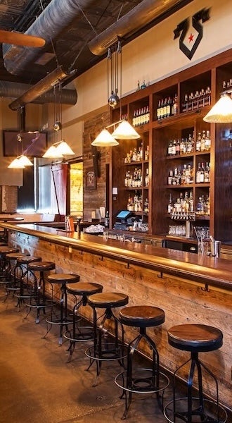 Bar interior with long wooden counter, stools and backlit shelves of bottles.