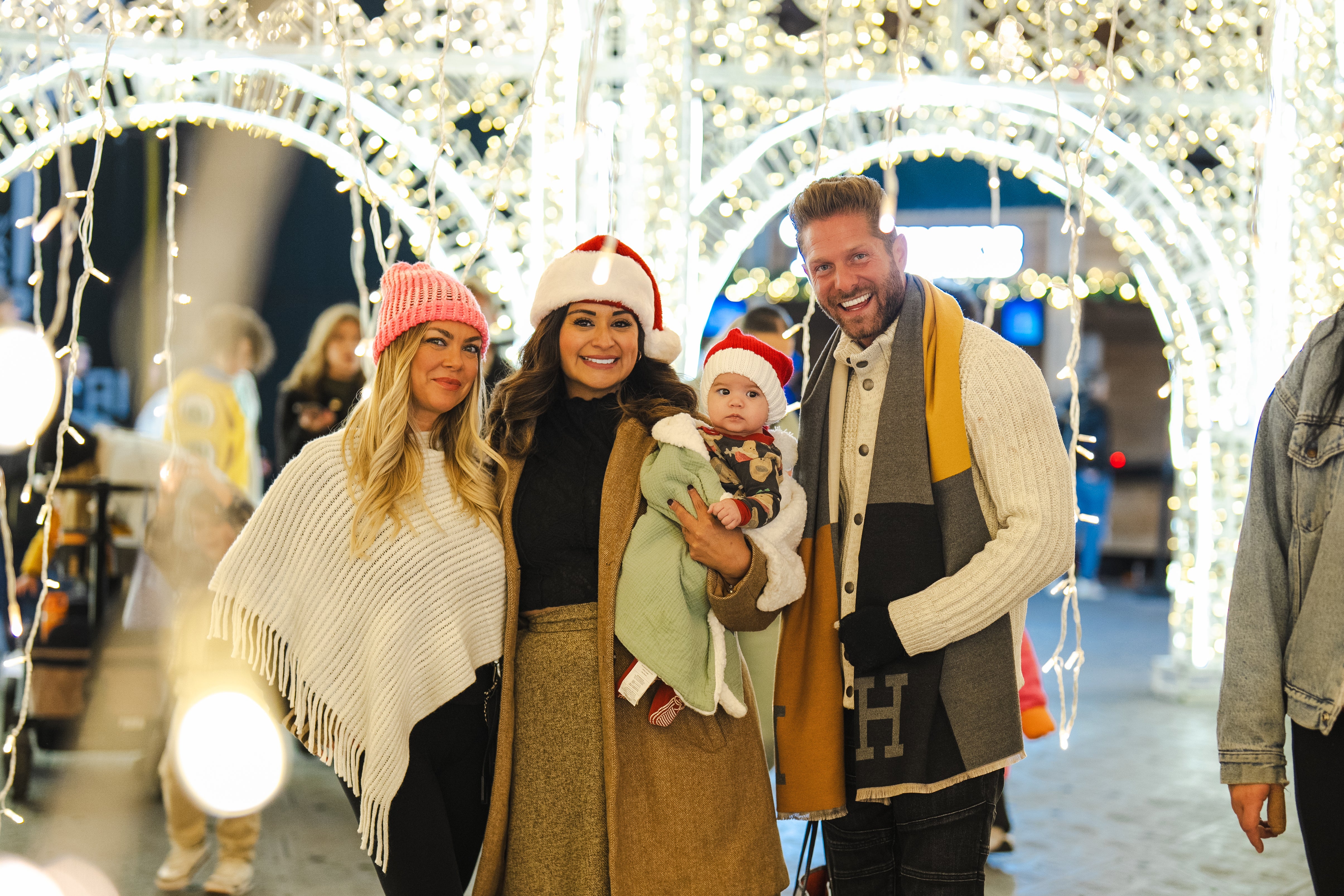 Photo: Three people in winter hats pose with small dog under festive light arch.