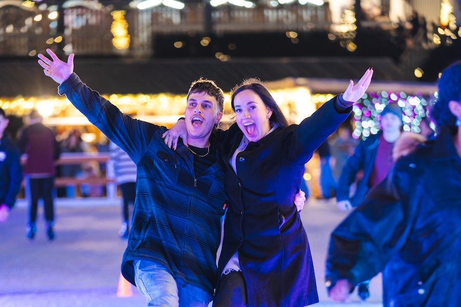 Photo: Two people ice skating at night with arms raised under bright rink lights