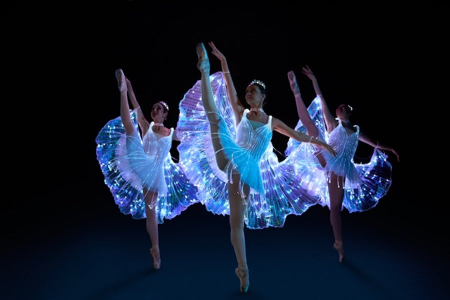 Ballerinas in shimmering blue costumes dance on a dark stage, arms raised