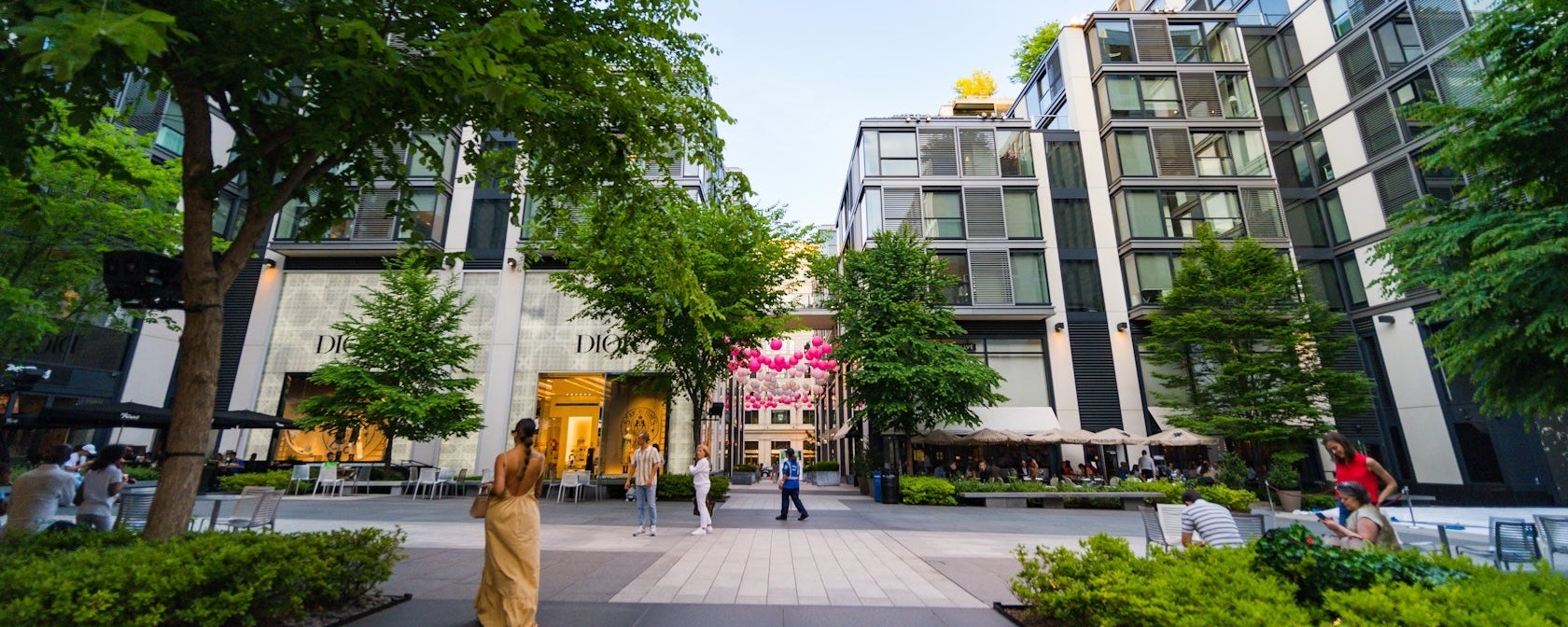 Modern glass building on tree-lined street with pedestrians and storefronts