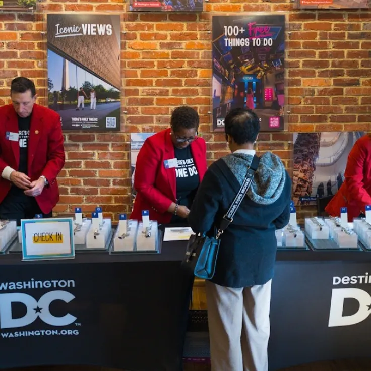 Destination DC Redcoat ambassadors assist attendees with event check-in at a registration table inside a brick-walled venue displaying Washington, DC promotional posters.