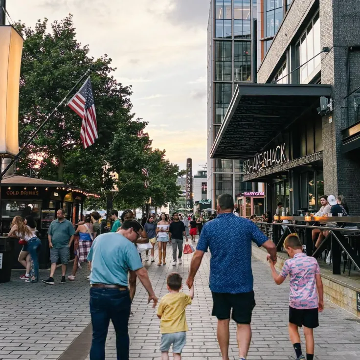 People walk on a DC street at dusk, with shops, U.S. flags and two children in front