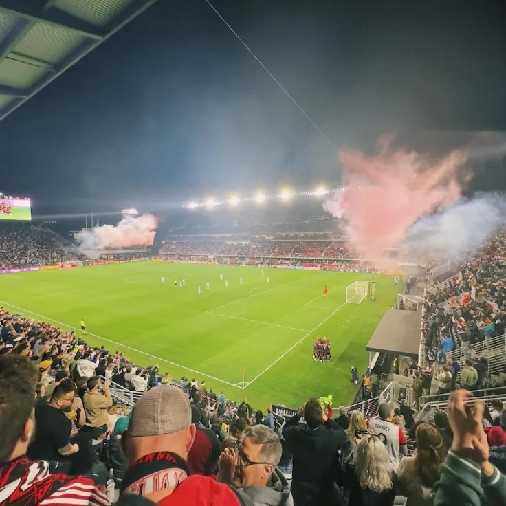 Photo of a packed stadium as pink smoke flares rise over the field at night