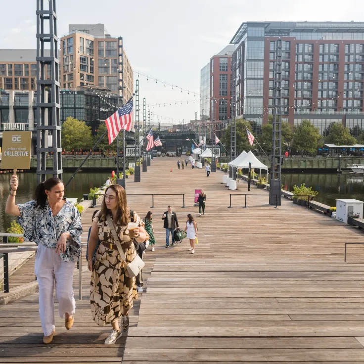 People stroll a wide wooden waterfront promenade lined with US flags and city buildings