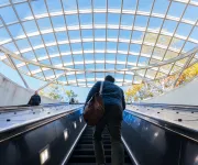 People ride up an escalator towards a skylight with a grid-like structure, revealing a bright blue sky and trees outside.