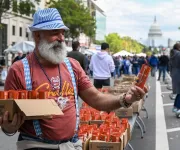A man with a white beard and a blue checkered hat and suspenders, handing out orange beer cans at an Oktoberfest event in Washington D.C., with the U.S. Capitol building visible in the background.