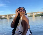A smiling woman wearing large sunglasses and a 'Bride to Be' sash poses near a river with an arched bridge in the background.