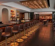 A stylish restaurant and bar with a long, sleek counter lined with leather barstools, illuminated liquor shelves, and a warmly lit dining area with modern decor in the background.