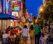 People walk down a street lit up with neon signs and busy restaurants and bars. 