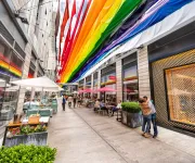 Colorful rainbow fabric banners hang above a lively pedestrian alley at CityCenterDC, where people dine outdoors and stroll between modern shops and restaurants.