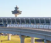 Silver Line Metro train traveling on elevated tracks with Washington Dulles International Airport terminal and air traffic control tower in the background.