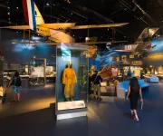 Visitors walk through an aviation exhibit with vintage aircraft and a yellow flight suit on display.