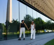 A couple holds hands while walking outside the Smithsonian National Museum of African American History and Culture, with the Washington Monument reflected in the glass.
