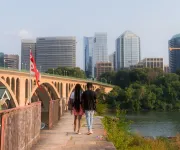 A couple walks along the Georgetown riverfront with a bridge and building of Arlington visible on the other side of the river. 