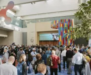 A large crowd of conference attendees gathers in a bright, spacious convention center lobby with colorful signage and modern architectural elements.