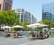 A lively outdoor plaza with people sitting under white umbrellas surrounded by modern glass buildings and leafy green trees on a sunny day.