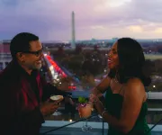 A couple enjoys cocktails on a rooftop terrace with the Washington Monument visible in the background at dusk.