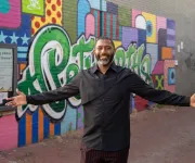 A smiling man stands with his arms open in front of a colorful street mural in Washington, DC.