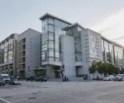 A modern glass-and-concrete museum building on a city corner with cars and pedestrians under a partly cloudy sky.