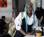 A person with long blue braids smiles while leaning on a café counter with people seated in the background.