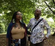 Two people in a leafy park, one holding a phone, both wearing patterned outfits.