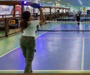 People playing pickleball on an indoor court with a net dividing the playing area and spectators visible in the background.