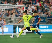 Photo of soccer player in yellow kicking toward goal, defended by blue-uniform player, packed crowd
