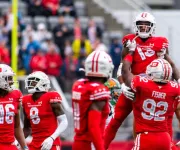 Photo, football players in red uniforms celebrate on the field as crowd watches.