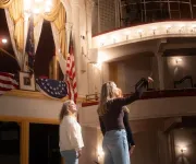Two performers on stage beneath ornate theater balconies and chandelier.