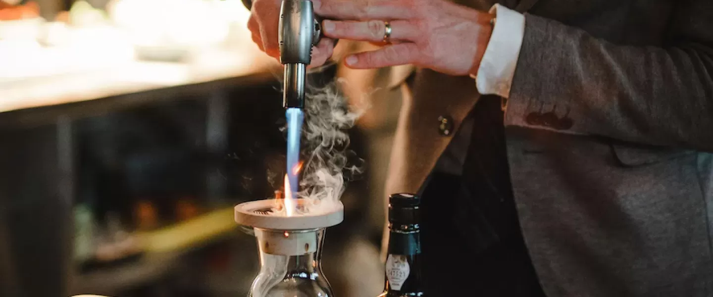 A person in a suit uses a torch to create a smoky effect in a glass decanter, with a bottle of liquor nearby.
