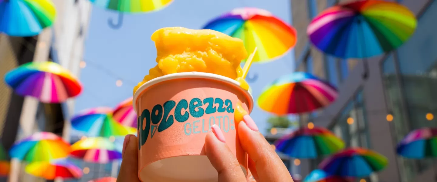 A hand holding a cup of orange gelato with vibrant, rainbow-colored umbrellas hanging overhead in the background.