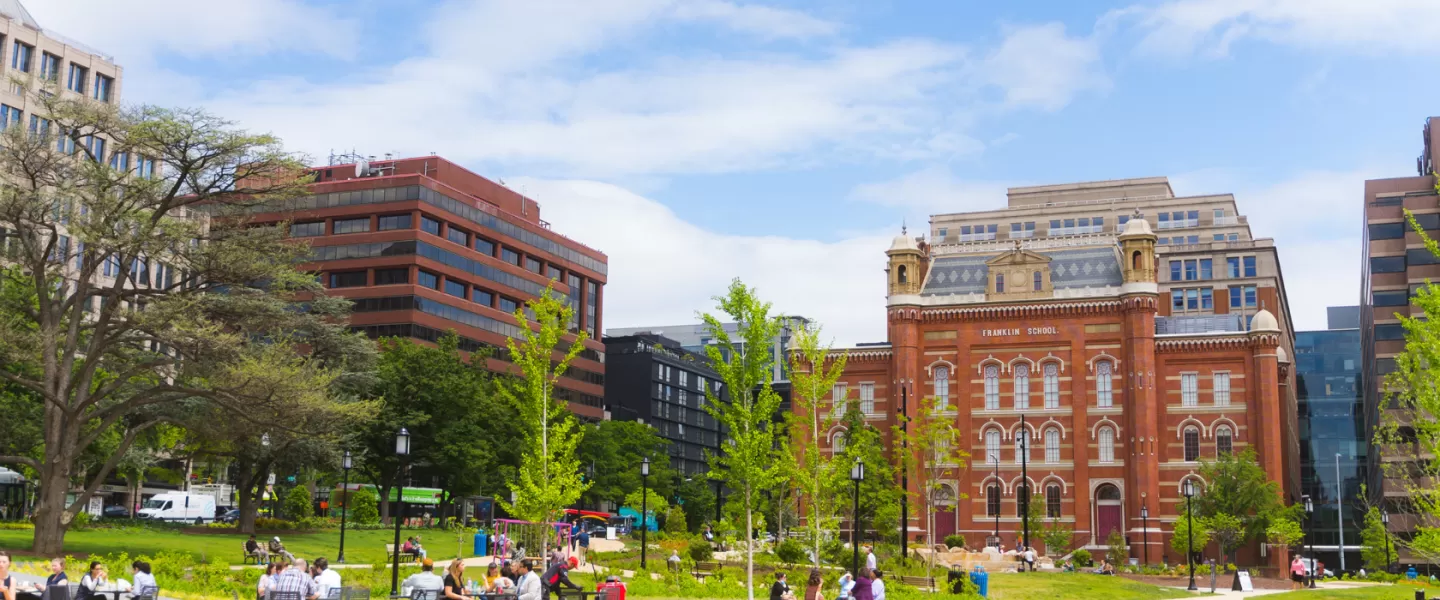 A sunny day in Franklin Square, Washington D.C., with people enjoying the park near the historic red-brick Franklin School building and surrounding office buildings.