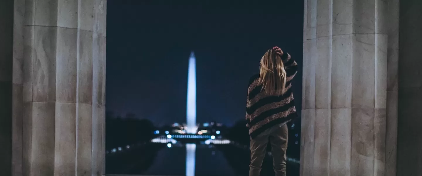 A photo from behind of a woman in a striped sweater looking at the Lincoln Memorial at night. 