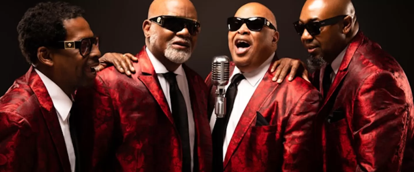 Four men, members of the Blind Boys of Alabama, are wearing matching red patterned suits and sunglasses. They stand close together around a vintage microphone, with one man singing while the others smile.