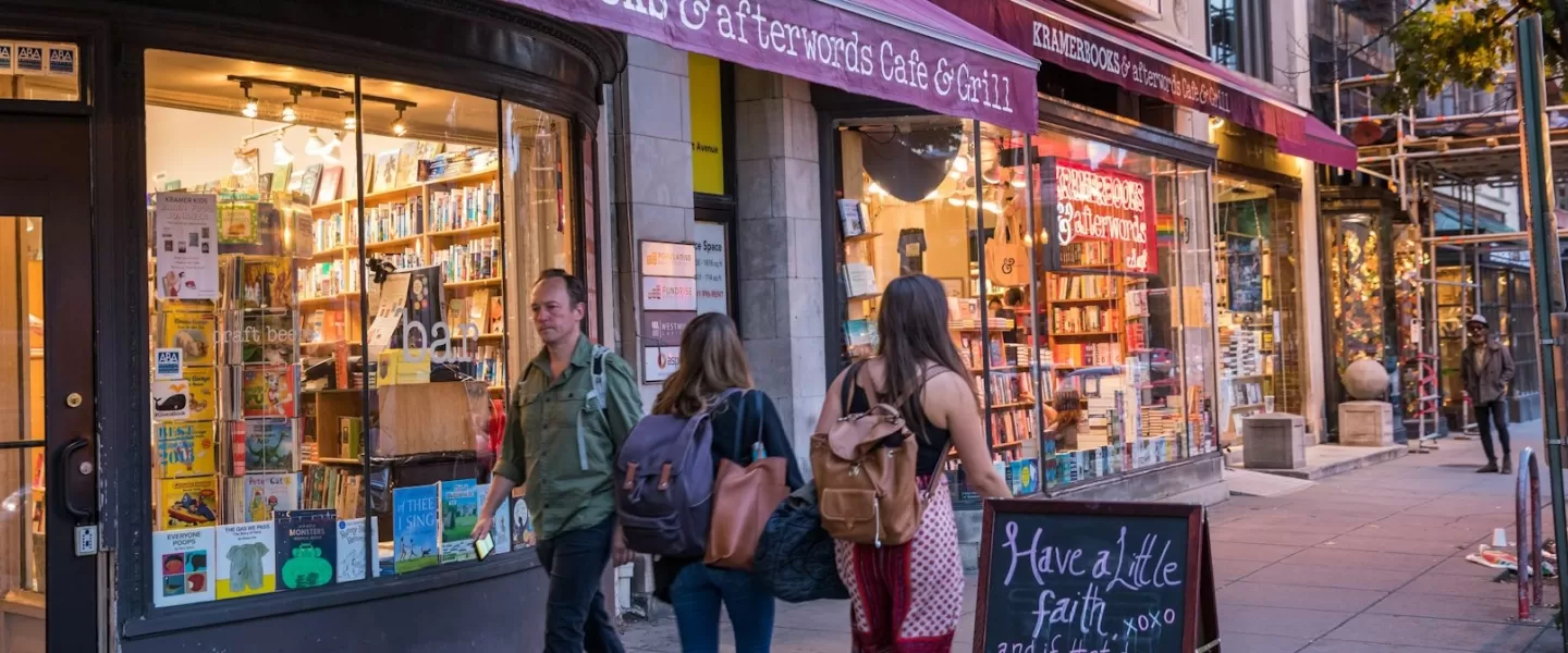 La gente pasea frente a la acogedora tienda Kramerbooks & Afterwords Cafe en Dupont Circle, con estantes de libros visibles a través de grandes ventanales y un cartel de pizarra en la acera.