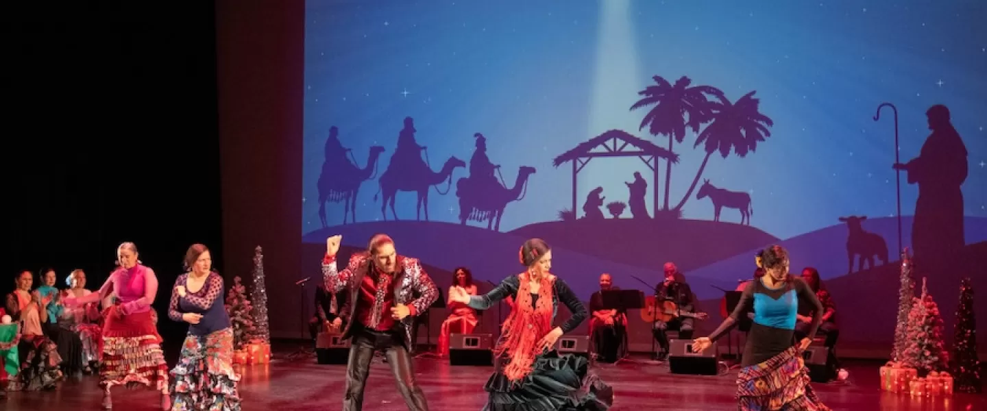 A group of dancers performing a vibrant flamenco routine on stage, with a nativity scene backdrop projected behind them. The dancers wear colorful, traditional outfits, and musicians are seated in the background.