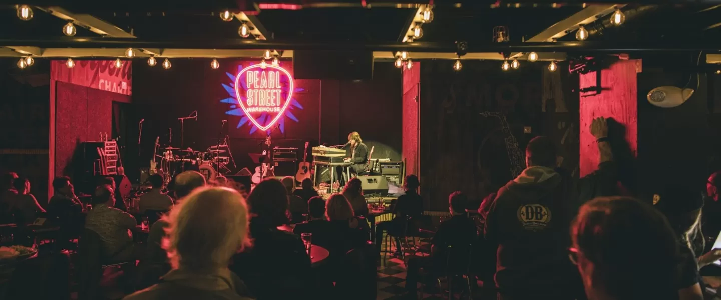 An intimate live music performance at Pearl Street Warehouse, with a neon sign in the shape of a guitar pick glowing on the back wall. The audience is seated, watching the performer on stage, with warm lighting overhead.