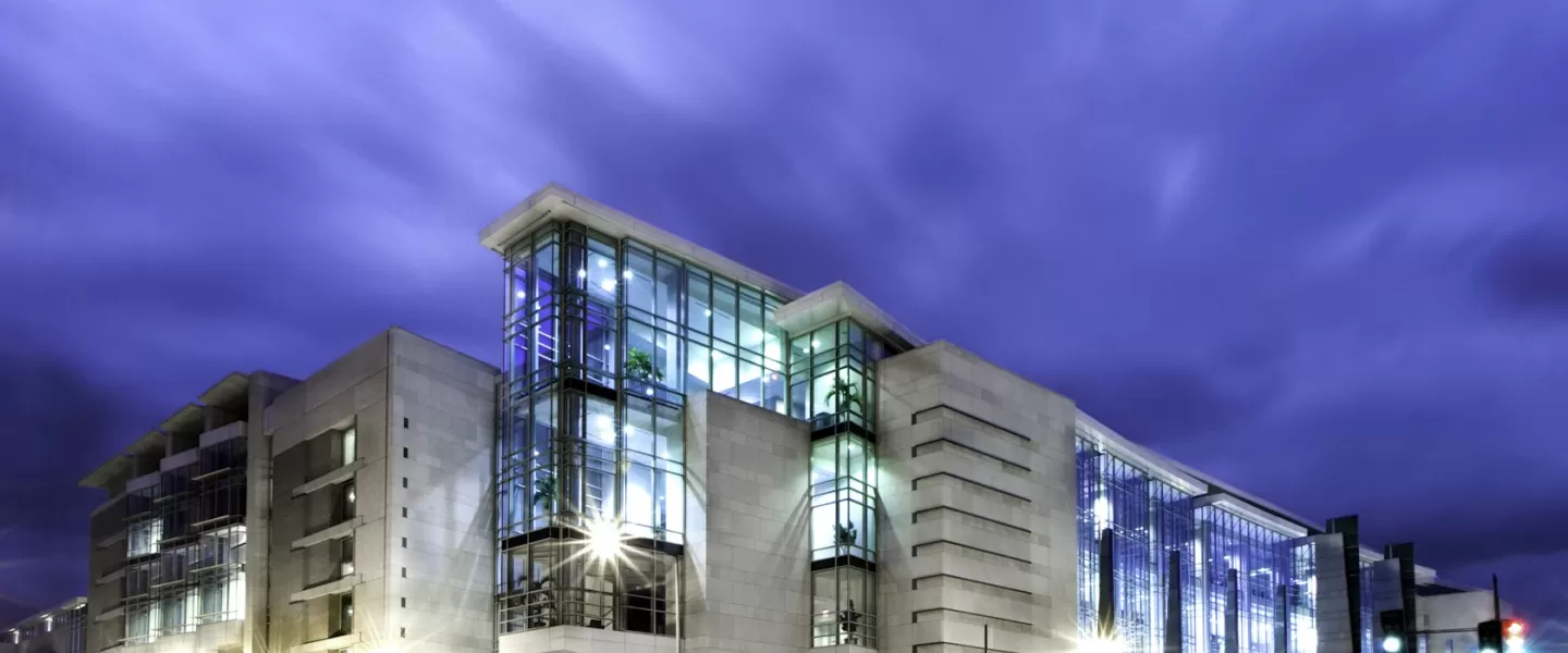 The image shows the exterior of the Walter E. Washington Convention Center in Washington, DC, illuminated at night. The sleek, modern architecture is highlighted by large glass windows and bright lighting, set against a dramatic sky. This convention center is a key venue for conferences, events, and exhibitions in the heart of the nation's capital.