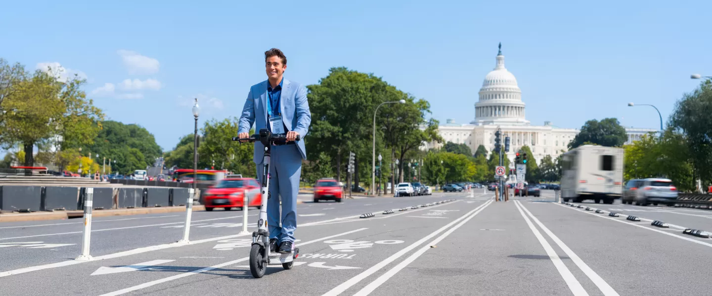 Un hombre con un traje azul conduce un scooter eléctrico por un carril bici con el edificio del Capitolio de Estados Unidos al fondo, disfrutando de un día soleado en Washington, DC.