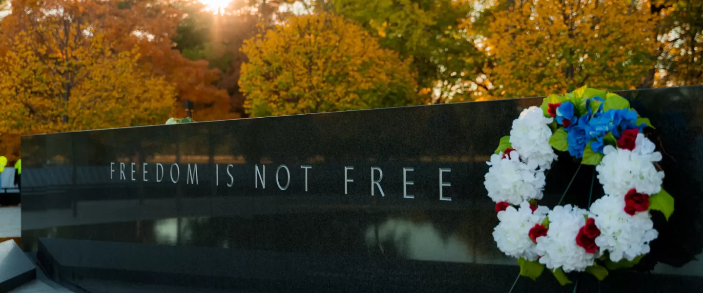 Una scena solenne presso un monumento commemorativo, con un muro di pietra su cui sono incise le parole "LA LIBERTÀ NON È GRATIS". Una corona di fiori rossi, bianchi e blu è appoggiata al muro, con alberi autunnali e il caldo bagliore del sole al tramonto sullo sfondo.