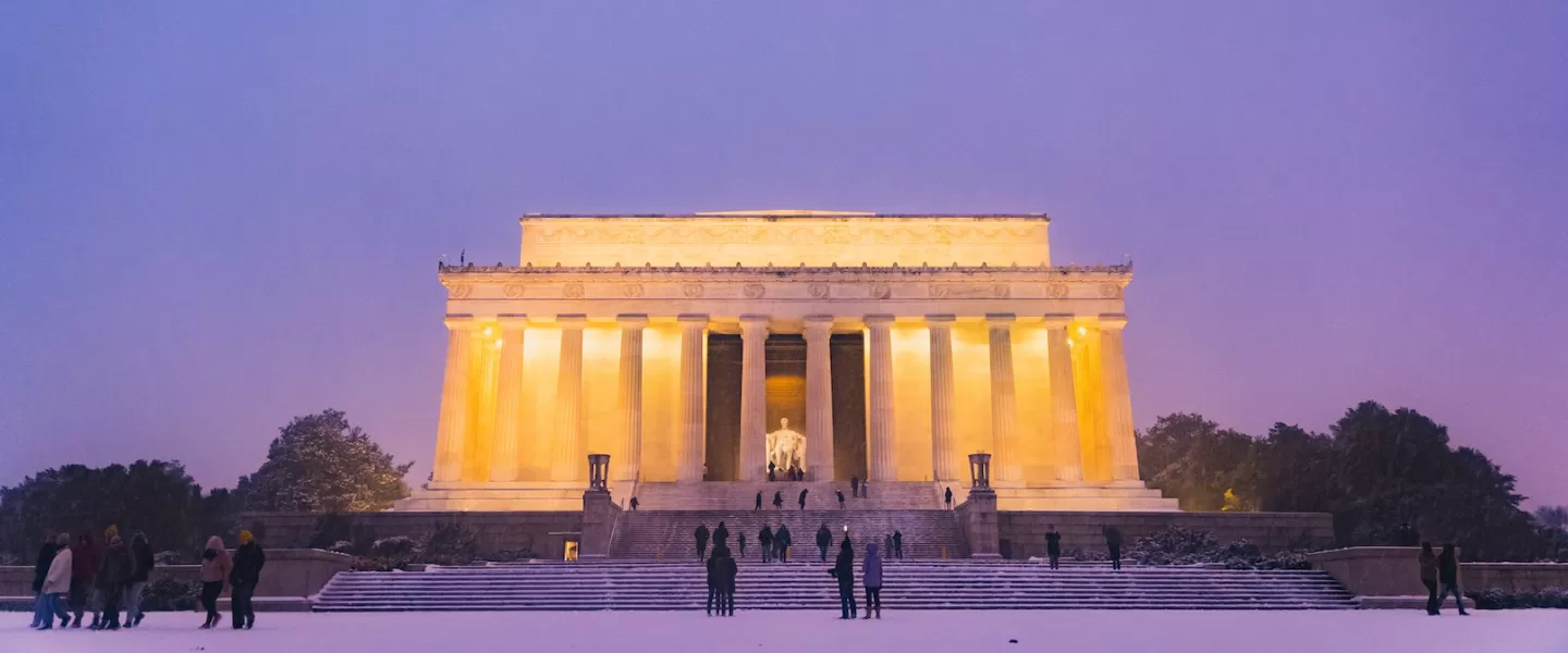 The Lincoln Memorial stands majestically against a twilight sky, illuminated in warm light. Snow blankets the ground as visitors stroll on the steps, with the iconic statue of Abraham Lincoln visible inside the memorial.