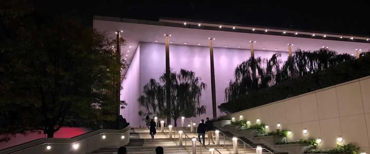 Nachtansicht der Außentreppe des Kennedy Centers. Menschen gehen hinauf und das Gebäude ist in sanftes violettes Licht getaucht.