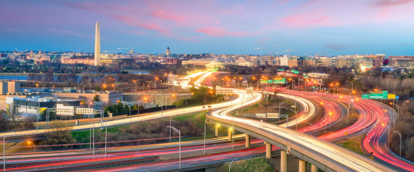  A vibrant view of Washington, DC, at twilight with light trails from vehicles on a highway interchange in the foreground and the Washington Monument standing tall in the cityscape.