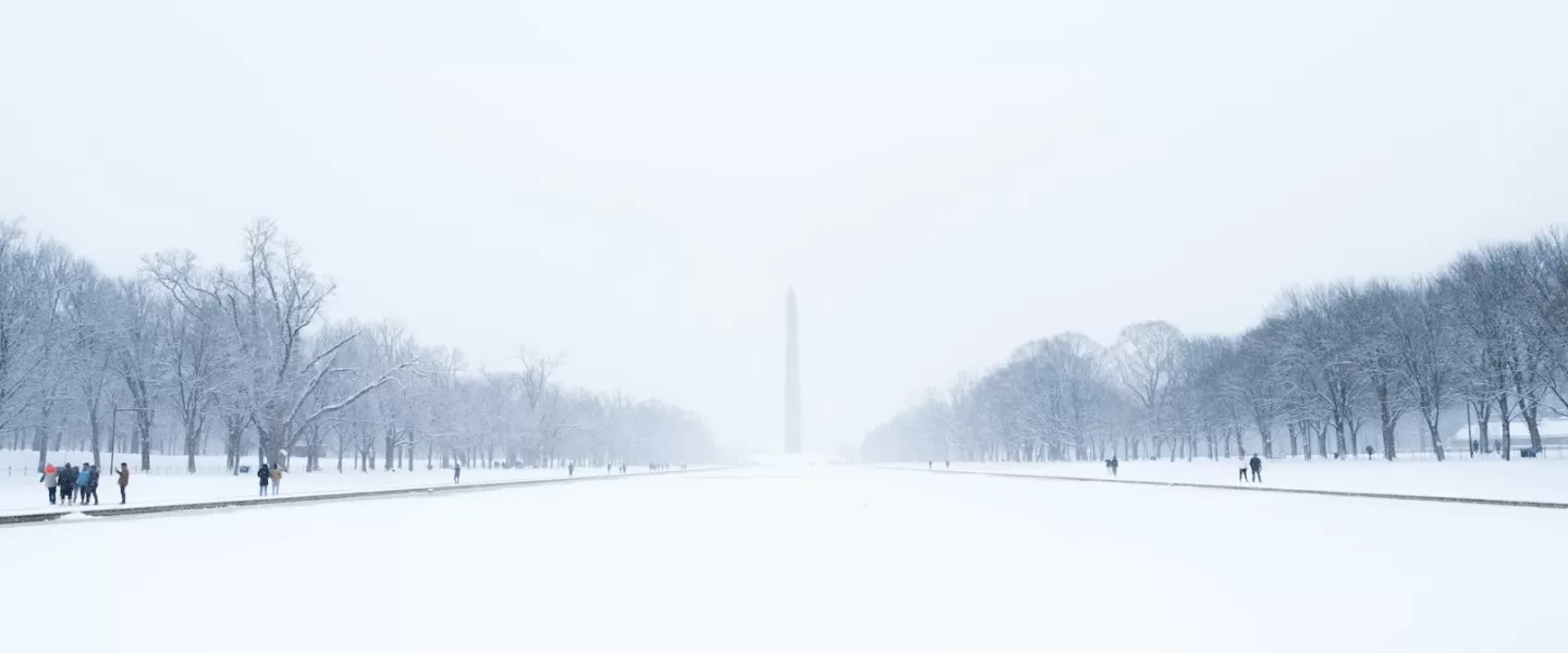  A snowy view of the Washington Monument framed by leafless trees and a snow-covered National Mall, with people strolling through the serene winter scene.