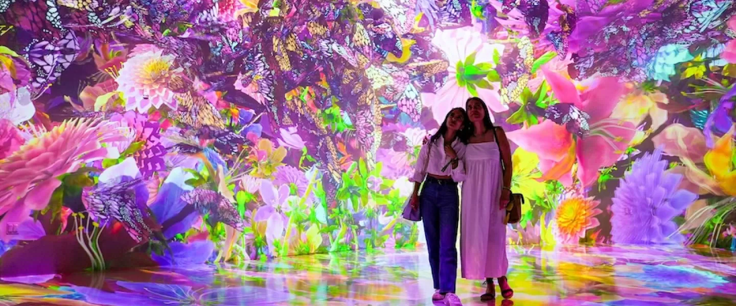 두 명의 방문객이 ARTECHOUSE의 생동감 넘치고 몰입감 넘치는 디지털 아트 설치물에 서 있는데, 그 주변은 다채롭고 빛나는 꽃과 나비로 둘러싸여 있습니다.